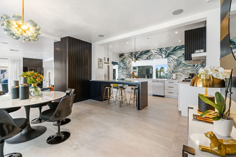 Bright open-plan kitchen and dining — tropical palm-leaf backsplash, dark island with gold bar stools, bubble chandelier, and white shaker cabinetry