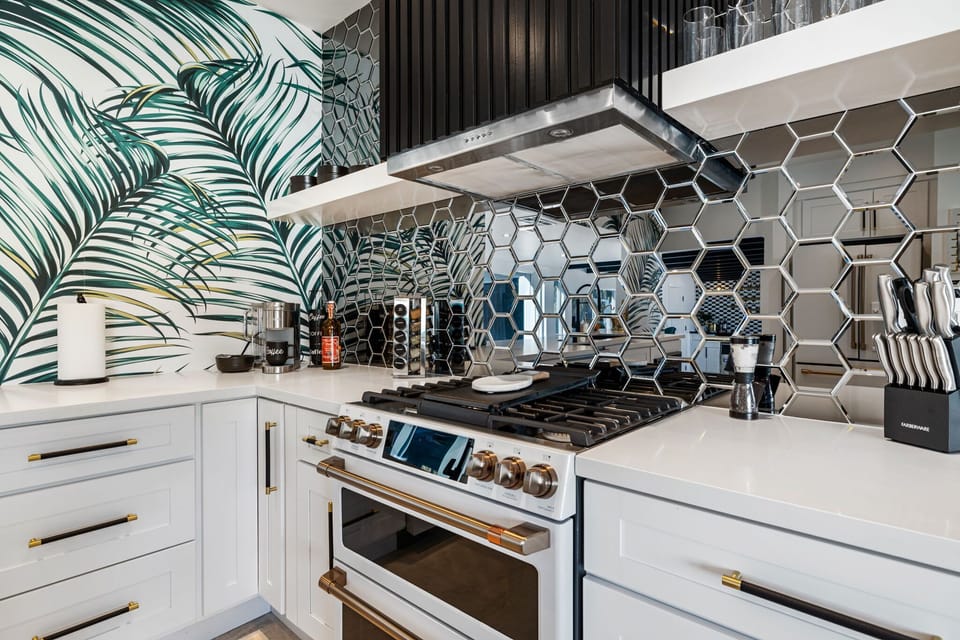 Kitchen closeup - pro gas range with gold hardware, mirrored hex tile backsplash, tropical palm-leaf wallpaper. Cooking here feels like a statement.