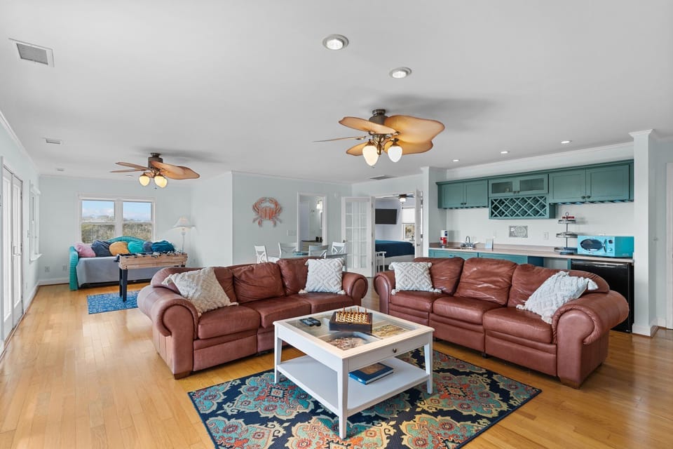 Spacious game room with leather sectional, TV, bookshelves, board games, wet bar, ceiling fan, coffee table, colorful rug, and wood floors.