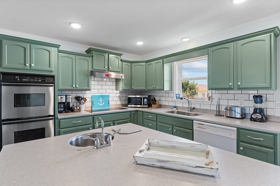 Coastal kitchen with green cabinets, center island sink, double oven, white tile backsplash, dishwasher, window, small appliances, and nautical decor.