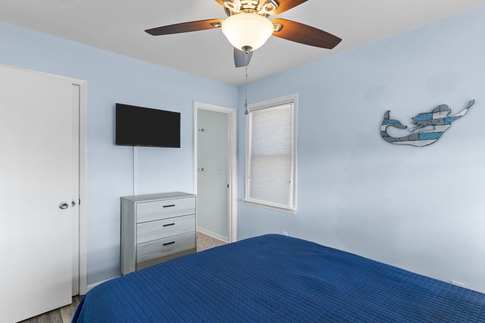 Spacious bedroom with king bed, blue bedding, ceiling fan, sitting chairs, side tables, carpet, light blue walls, and private bathroom access.