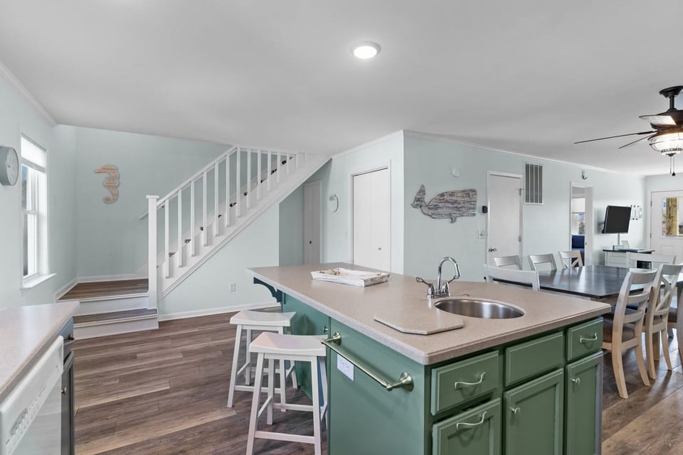 Coastal kitchen with green cabinets, center island sink, double oven, white tile backsplash, dishwasher, window, small appliances, and nautical decor.
