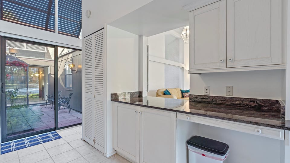A kitchen area with white cabinets, a granite countertop, and a trash bin. A sliding glass door opens to an outdoor patio with seating, visible through the doorway.