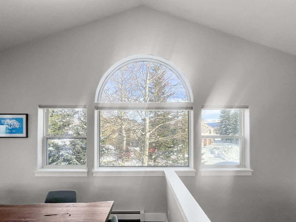 A living space with a high ceiling features a large arched window with a view of snow-covered trees and houses outside. Light streams through the window, illuminating the room.