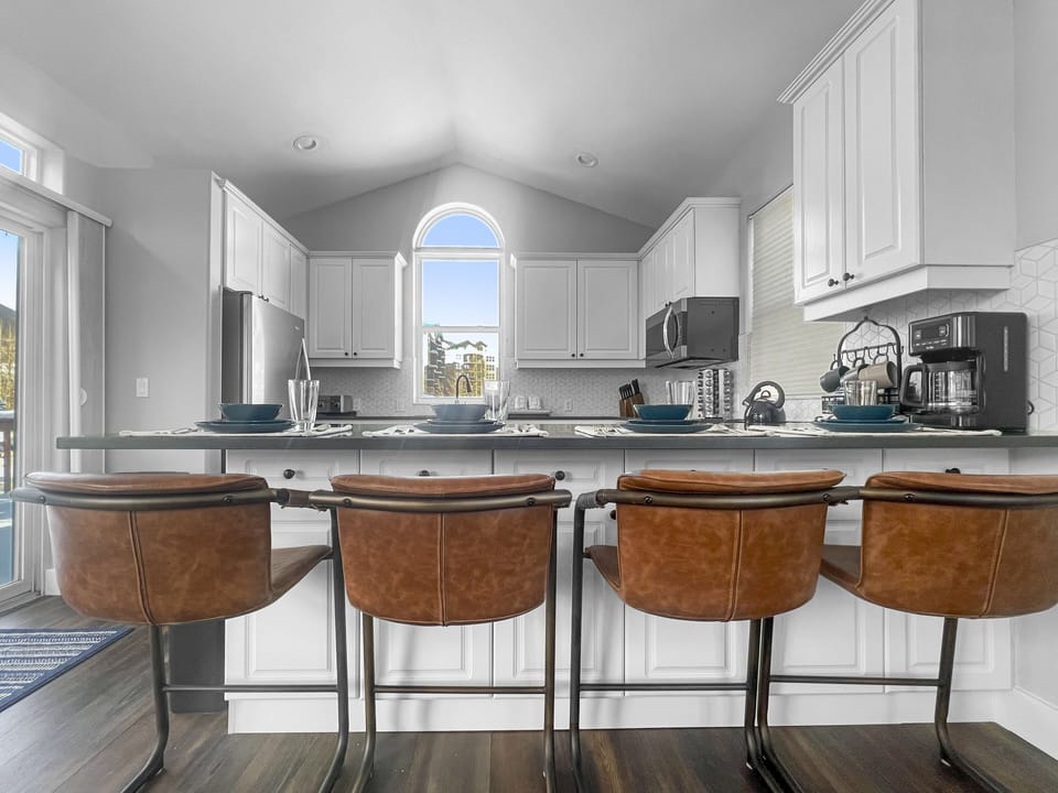 A modern kitchen with white cabinets, stainless steel appliances, a central island with brown leather bar stools, and a high arched window bringing in natural light.