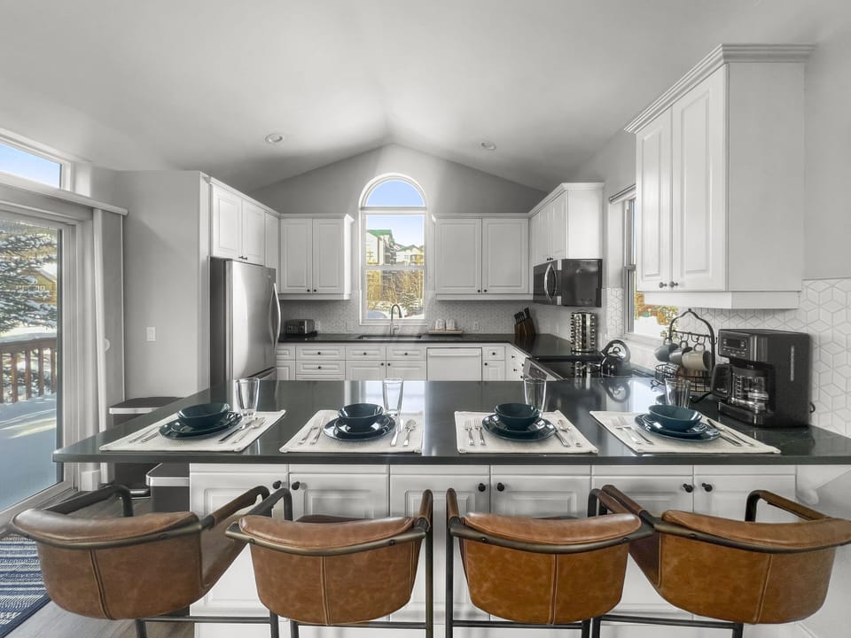 A modern kitchen with a center island, four barstools, white cabinetry, stainless steel appliances, and a large arched window above the sink.