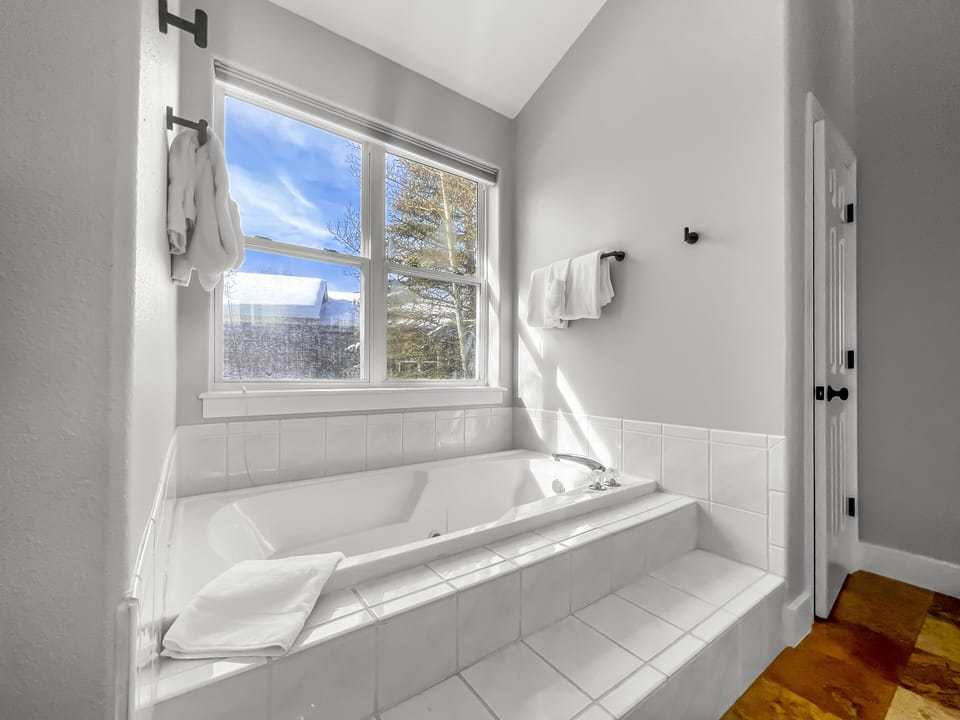 A white tiled bathtub with a towel on the edge sits by a large window with a view of trees and a blue sky. A towel rack with white towels is mounted on the wall beside the window.
