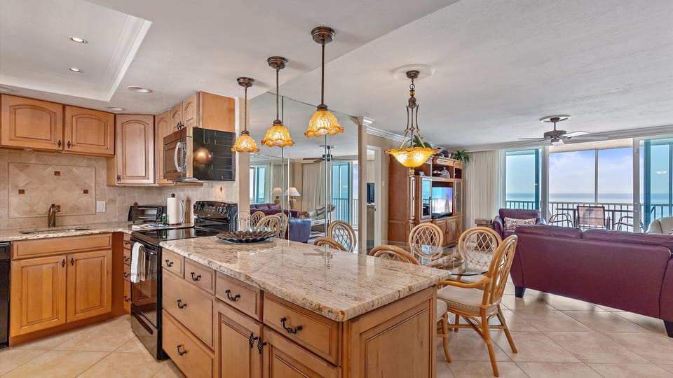 A modern open-plan kitchen and living area with a granite-topped island, wooden cabinets, a dining table, and a sitting area. Large windows provide a view of the ocean from the balcony.