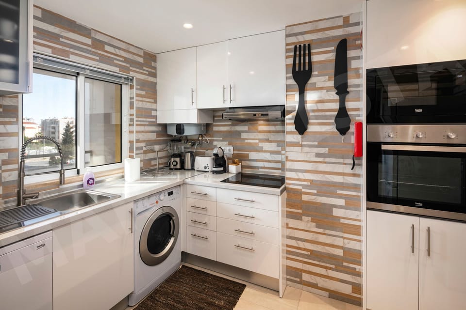 Modern fully equipped kitchen in Apartment Mar a Vista with stylish wood-effect tiled splashback, white gloss cabinets, built-in oven, washing machine, and quirky fork and knife wall decor, perfect for self-catering in the Algarve