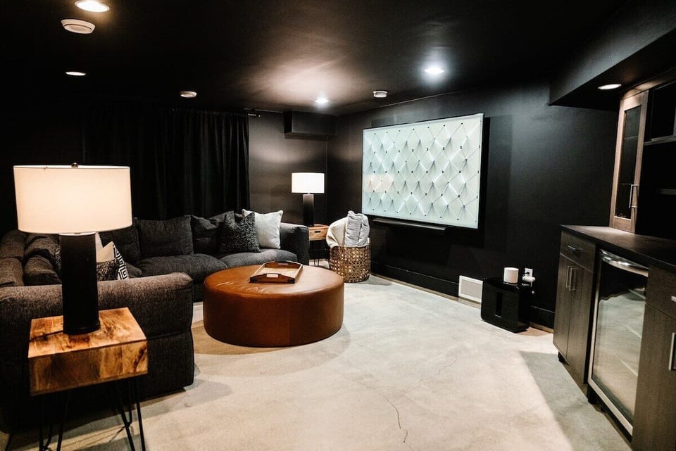 This modern basement living room offers a cozy, cinematic vibe with dark charcoal walls and ceiling. A large sectional sofa, brown leather ottoman, and sleek wooden bar area set the mood for relaxing or entertaining.