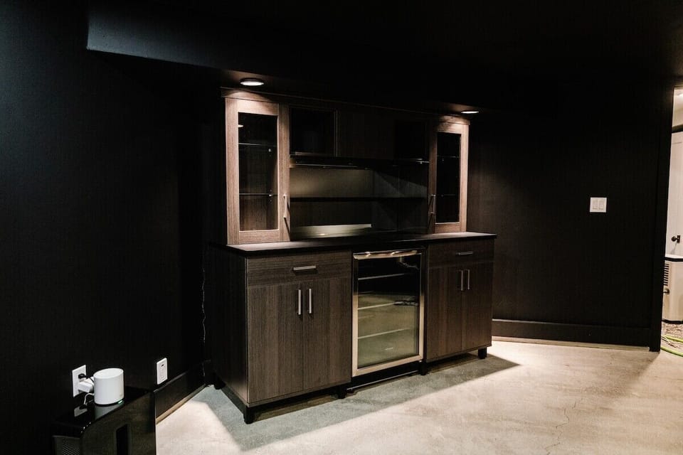 This stylish home bar area invites guests to enjoy a drink, with a light gray wooden bar cabinet featuring glass shelves and a beverage fridge. Relax with a drink from the black side table and mugs, or unwind in the cozy corner.