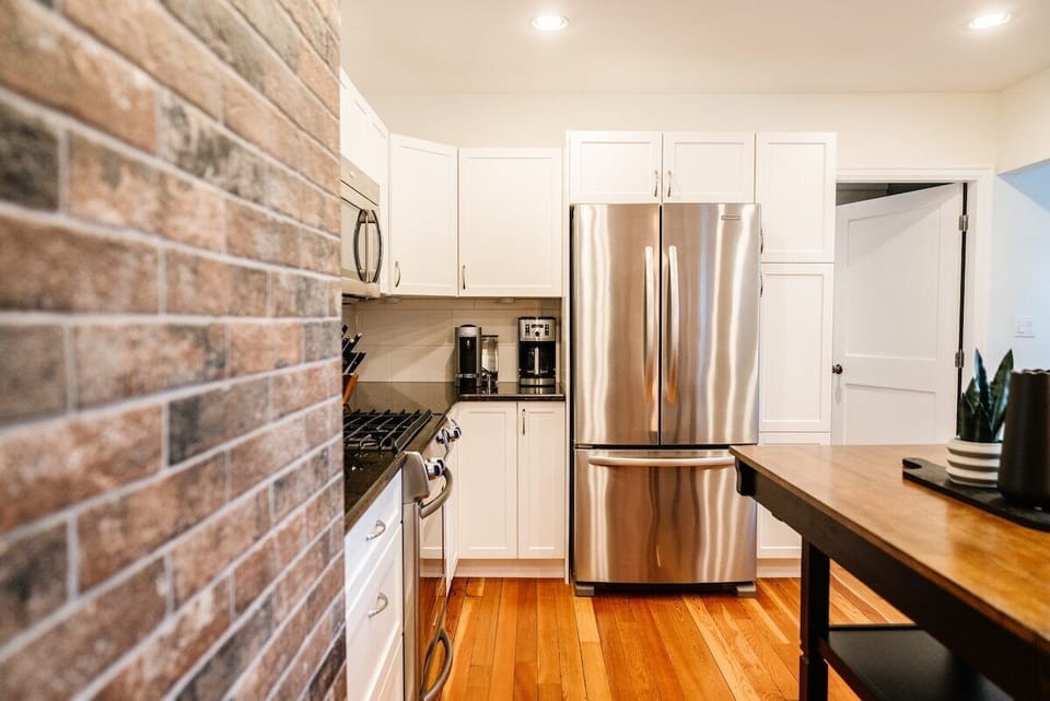 This modern kitchen boasts stainless steel appliances, white cabinets, and a wood floor. A brick wall adds texture, while a potted plant on the wooden table enhances the clean, well-lit space.