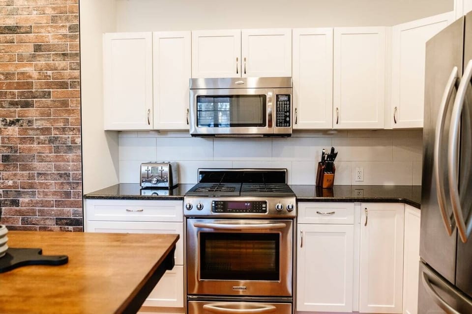 This kitchen combines style and functionality with white cabinets, black granite countertops, and stainless steel appliances. Prepare meals with ease using the toaster and knife block, all set against a warm brick backsplash