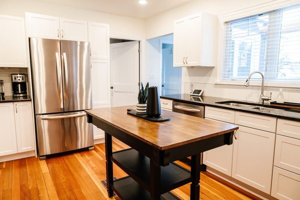This bright kitchen exudes warmth with its white cabinetry, dark countertops, and wood flooring. The butcher block island, adorned with potted plants and a tablet, offers a cozy spot to gather, while natural light fills the space.