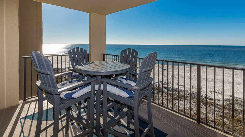 The balcony features a round table with tall chairs, offering a serene view of the tranquil beachfront and shimmering water