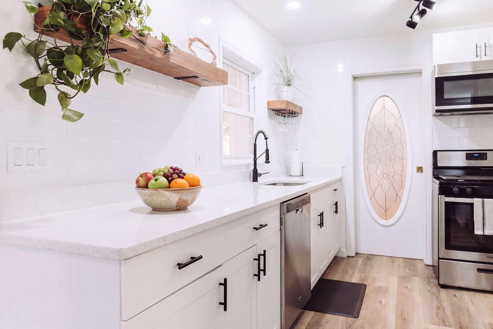 Prepare your favorite meals in this bright, airy kitchen featuring crisp white cabinetry, stainless steel appliances, and elegant quartz countertops. The open shelving with lush greenery adds a touch of warmth to this high-end, functional space.