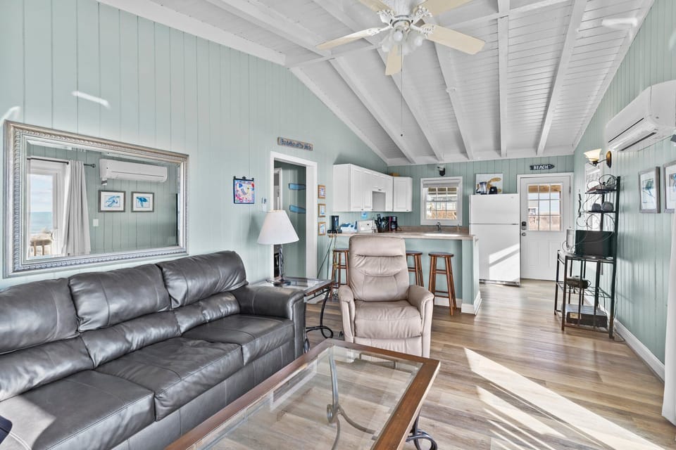 Bright living room with sofa, recliner, glass coffee table, vaulted ceiling, kitchen, sliding doors, deck chairs, dunes, and ocean views in Oak Island, NC.