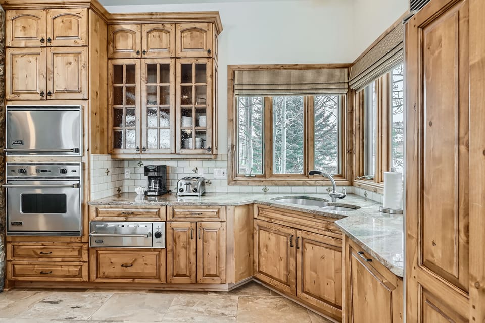 A kitchen with wooden cabinets, a double oven, a stove, a coffee maker, a toaster, and a sink. Large windows provide natural light and views of a snowy landscape outside.