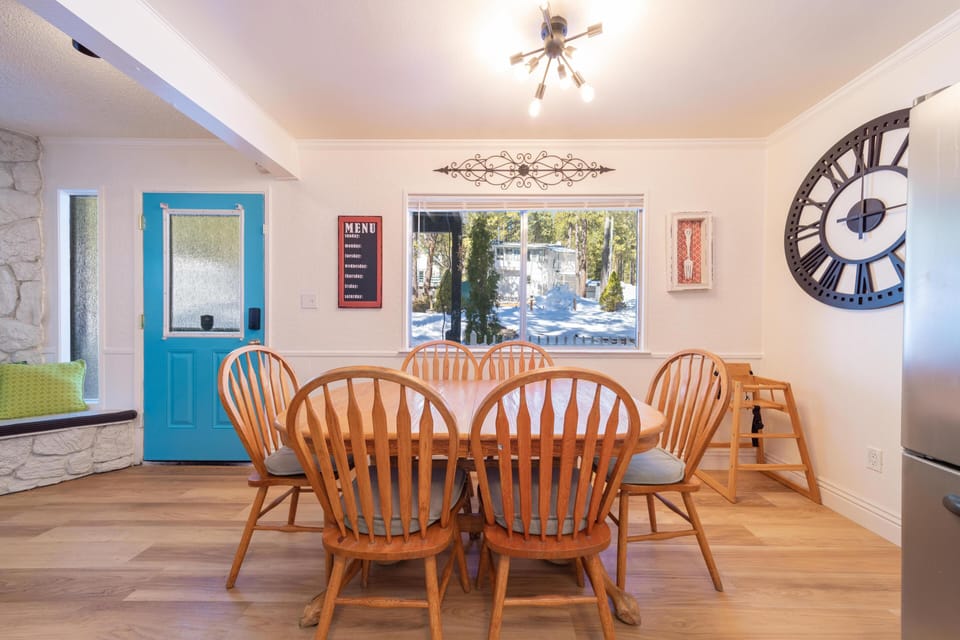 Wood dining table with six spindle-back chairs sits beneath a modern sputnik light and large wall clock.