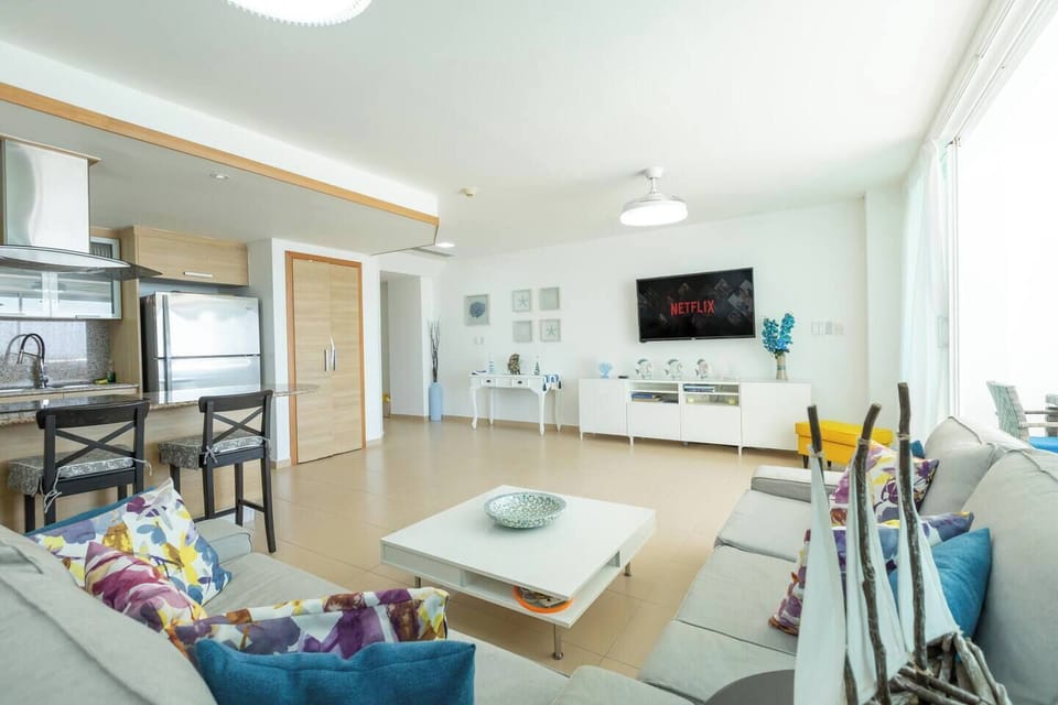 Spacious living room with a 55-inch Smart TV, A/C, and open layout—connected to the kitchen on one side and the balcony with ocean views on the other.