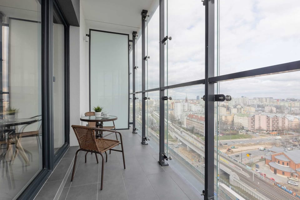 A cozy balcony with glass railings, perfect for enjoying the surrounding city view. A small table adds functionality to the space.