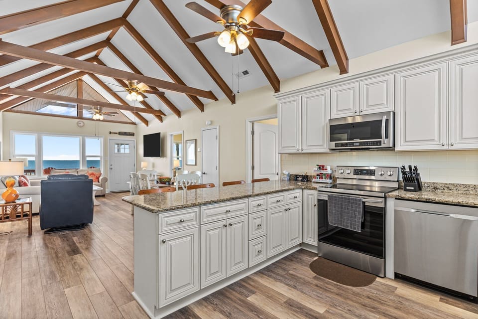 Open kitchen with granite island seating for four, white cabinets, stainless appliances, vaulted ceiling with exposed beams, ceiling fan, and large windows bringing in natural light.
