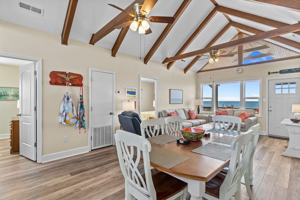 Coastal living and dining space with vaulted wood-beam ceilings, dining table for six, comfortable seating, and large windows showcasing direct ocean views and natural light.