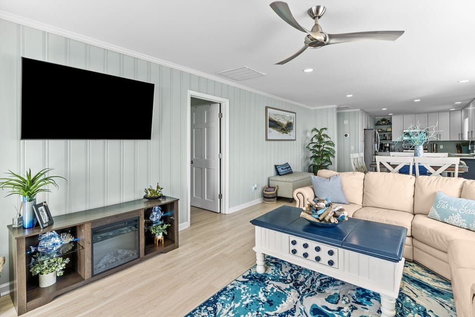 Bright Oak Island oceanfront living room with leather sofas, coastal rug, ceiling fan, wall-mounted TV, and sliding glass doors to a deck with turquoise chairs. Adjacent bedroom with blue bedding visible just off the main space.