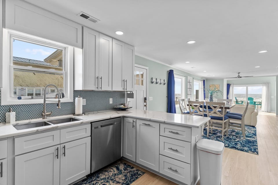 : Light-filled Oak Island kitchen featuring white shaker cabinets, quartz countertops, stainless steel dishwasher, double sink under window, mosaic tile backsplash, and open sightlines to the dining area and oceanfront deck beyond.