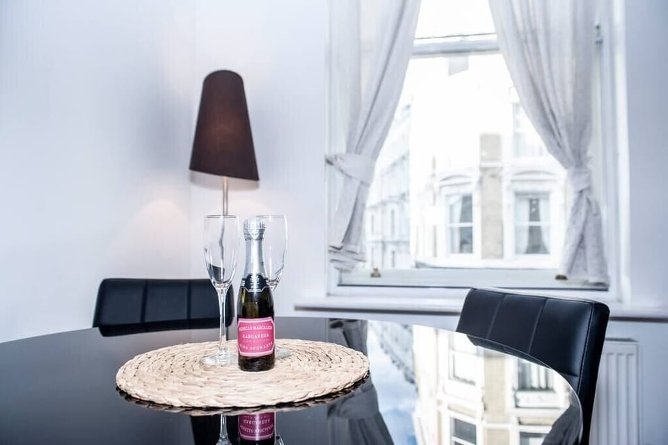 The spacious, bright living room with high ceilings creates an open, airy atmosphere. It's furnished with a comfortable sofa and armchair, perfect for lounging, and a sleek glass round table with chairs, providing a stylish and functional space.
