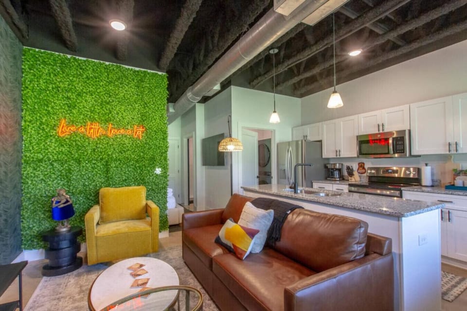 Open-concept kitchen and living area with a green moss accent wall, glowing neon signs, and stainless appliances