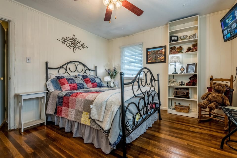 Iron-framed bed with patchwork quilt stands on polished hardwood floors beside a white bookshelf and rocking chair.