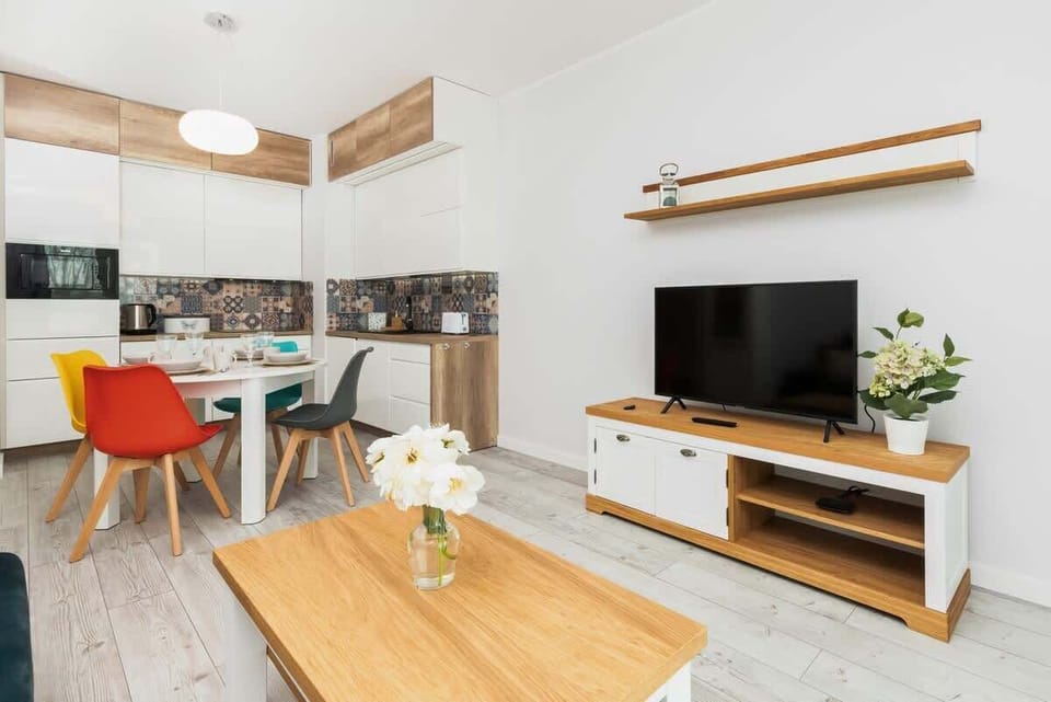 A living room with a TV mounted on the wall, a wooden coffee table, and an adjacent dining area with vibrant orange chairs.
