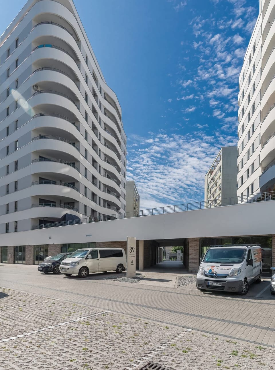 An exterior shot of a modern apartment building with a curved facade, balconies, and a clean paved walkway in the foreground. The sky is clear and blue.