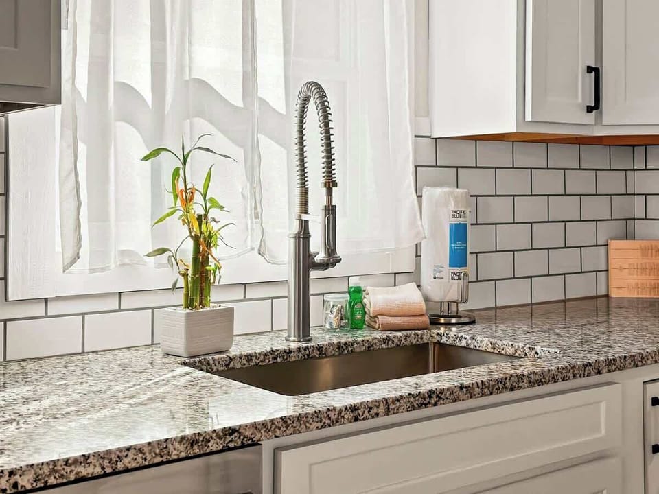 A culinary oasis: a kitchen sink adorned with a convenient pull-down faucet, overlooking windows dressed in white curtains, complemented by a lucky bamboo plant for a touch of natural charm.