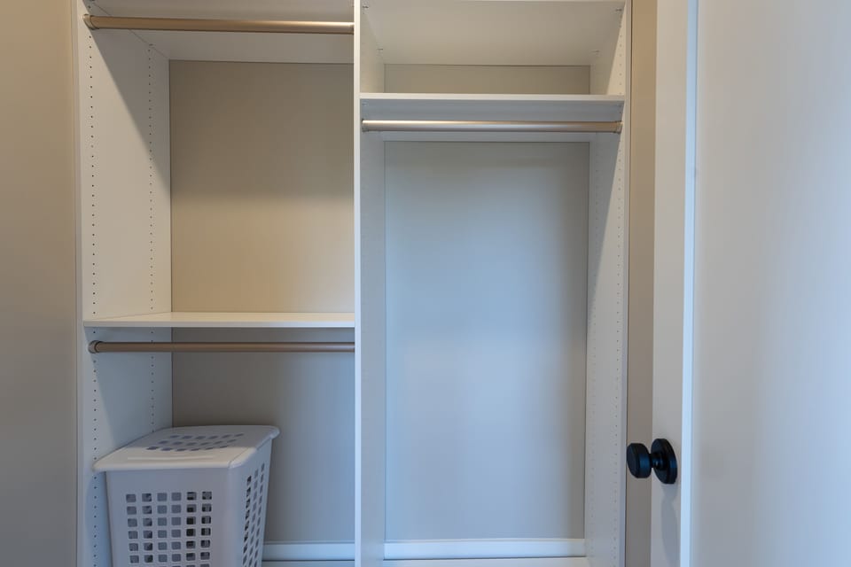 Empty white closet with open door, two hanging rods, shelving, and a laundry basket on the lower left.