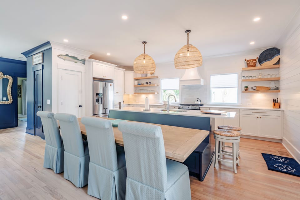 Four light blue slip covered parson's chairs surround a large rectangular kitchen table on one side and a wooden bench seat on the far side of the table. The bench seat is connected to the kitchen island which is painted in Navy blue.