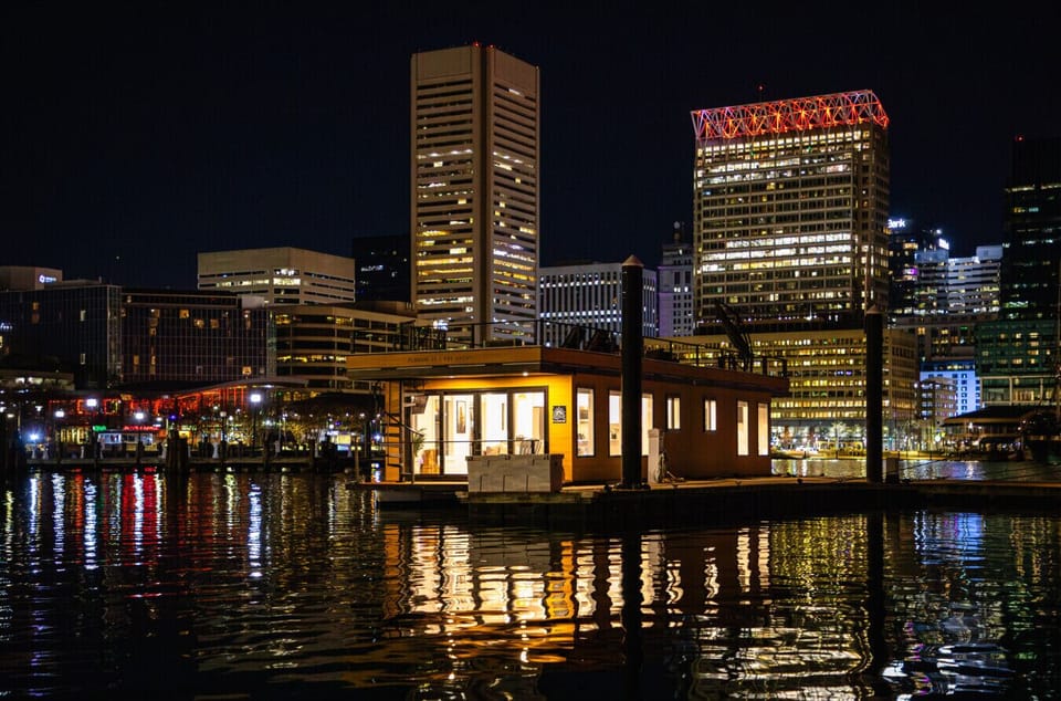 Bay Escape glowing from across the Inner Harbor at night, with the Baltimore World Trade Center and downtown skyline towering behind.