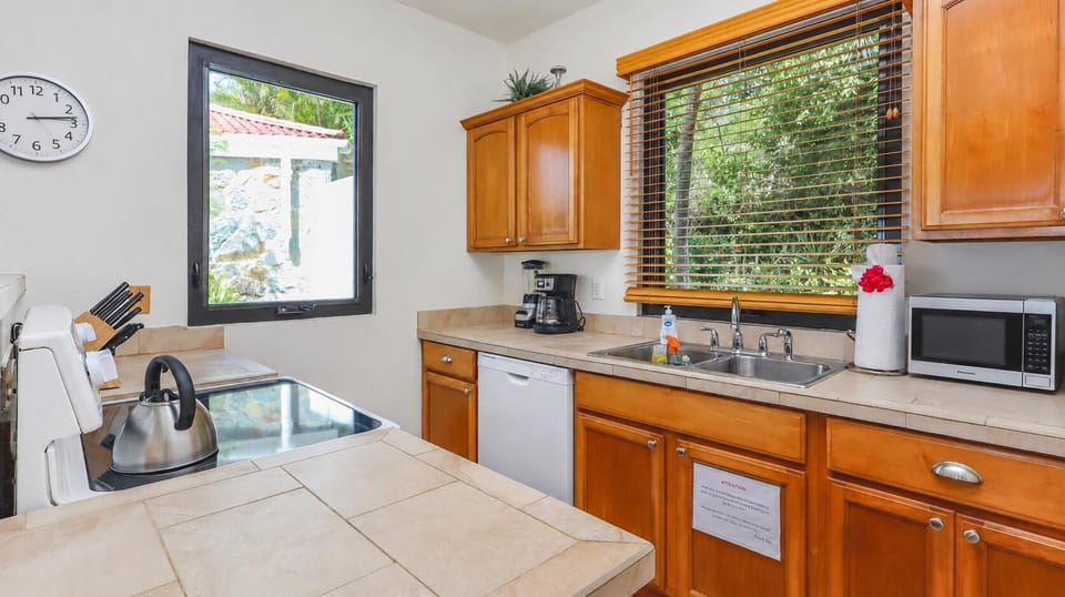 Tile countertops surround a double sink under wooden blinds with leafy green views in the kitchen.