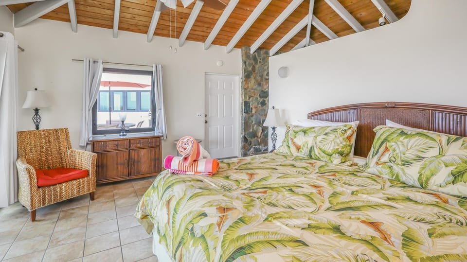 King bed with tropical leaf bedding sits beneath a vaulted wood ceiling next to a wicker armchair and dresser.