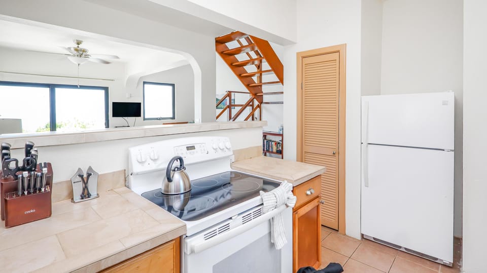 White electric stove with stainless steel kettle sits between beige tile countertops and light wood cabinets.