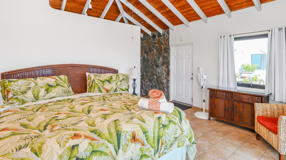 King bed with tropical-print bedding sits beneath a vaulted wood ceiling and beside a natural stone accent wall.