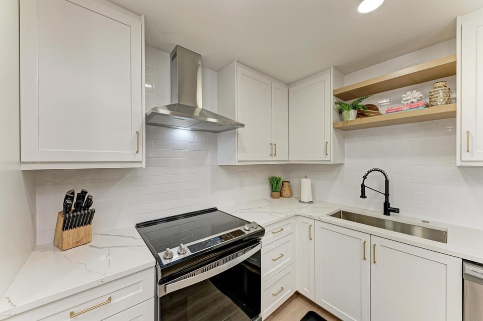 Open kitchen design with sleek oven and stove. Metal vent hood for cooking. Sink with shelving above