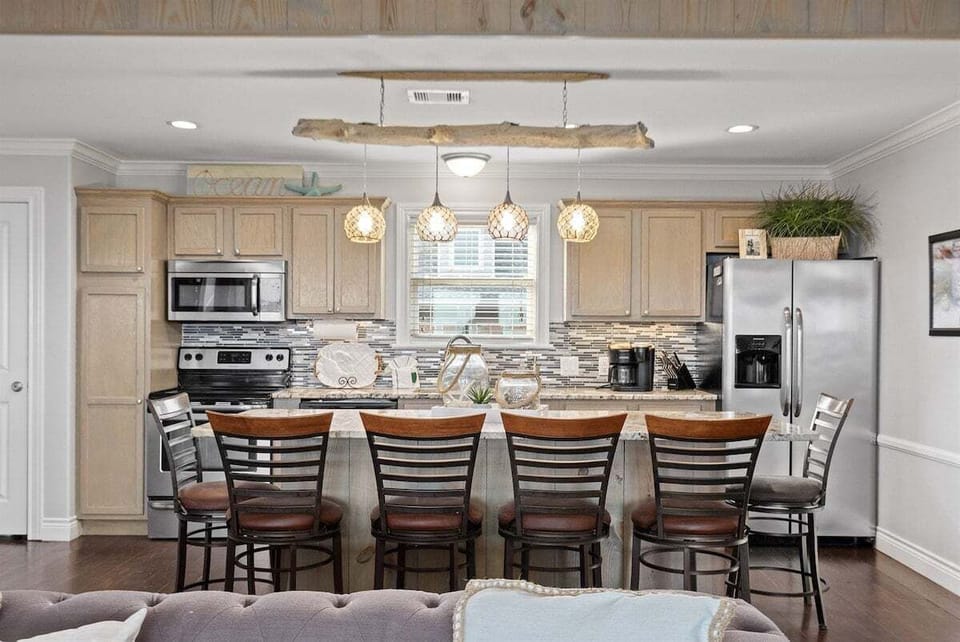 Open kitchen with breakfast bar seating, stainless steel refrigerator, range, microwave, pendant lighting, and tile backsplash.