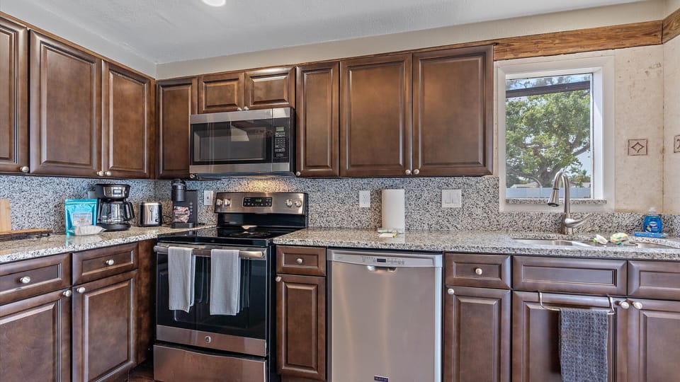 Modern kitchen with dark wood cabinets, granite countertops, stainless steel appliances, a microwave, oven, dishwasher, and a window above the sink. Coffee maker and kitchen essentials on the counter.