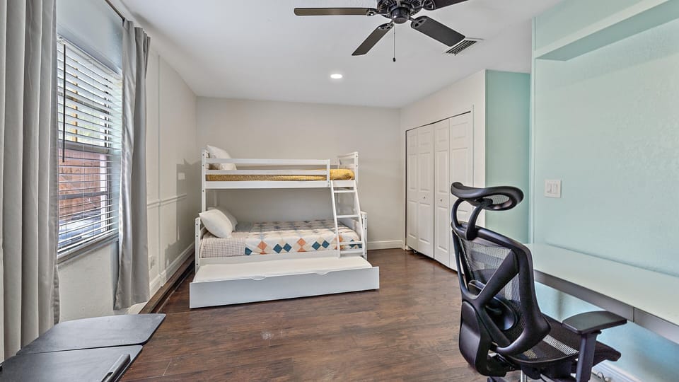 A bedroom with wooden floors features a bunk bed, a desk with a chair, a ceiling fan, and a window with closed curtains.