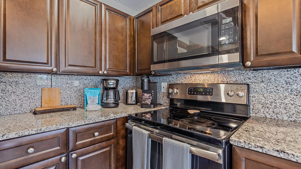 A modern kitchen with dark wooden cabinets, granite countertops, a stainless steel microwave, and an electric stove. Various kitchen items including a coffee maker, toaster, and cutting board are placed on the counter.