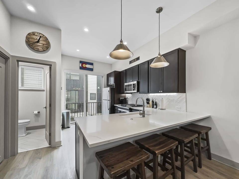 Modern kitchen with dark wood cabinets, white countertops, and three wooden barstools at the island. Hanging pendant lights, stainless steel appliances, and a view into a small bathroom and balcony.