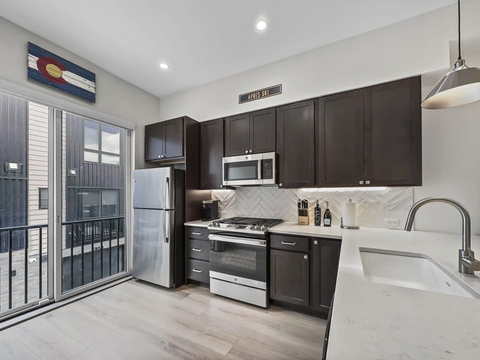 Modern kitchen with stainless steel appliances, dark wood cabinets, white herringbone backsplash, and a kitchen island. A large window on the left provides natural light and a balcony view.
