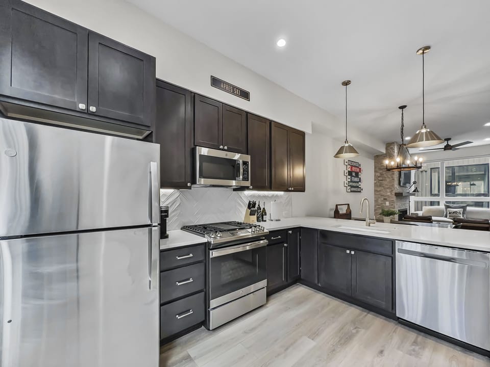 Modern kitchen with stainless steel appliances, dark cabinets, a gas stove, overhead microwave, and a dual sink. Wooden flooring and a view into the dining and living areas.